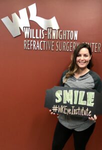 Patient posing for a photo in front of the "Willis Knighton Refractive Surgery Center. They're holding a sign that reads"SMILE #WKEyeInstitute"