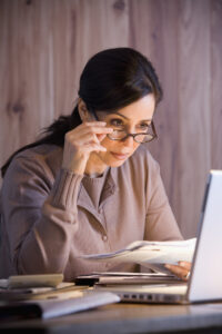 A woman working on her computer, while holding papers and tilting her glasses